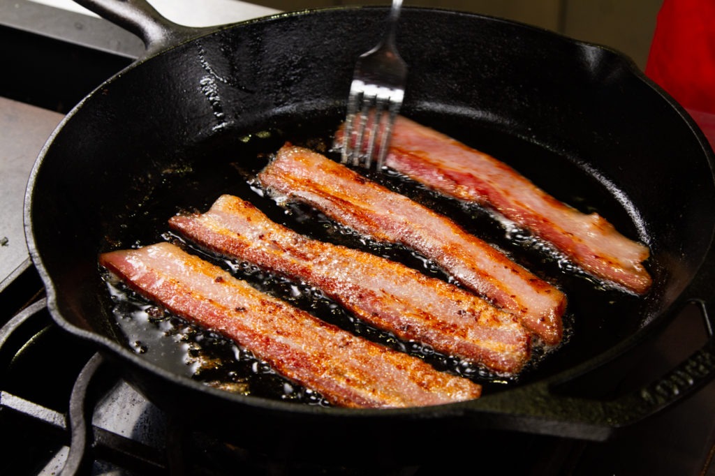 Home cured bacon frying in a cast iron skillet.