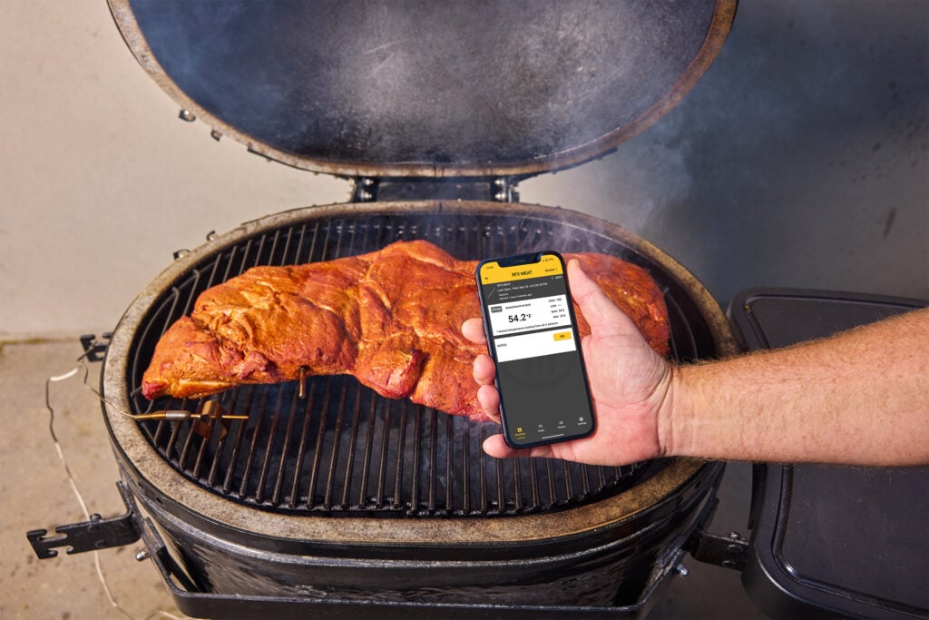 ThermoWorks app shown in front of butterflied pork butt on a kamado-style smoker grill