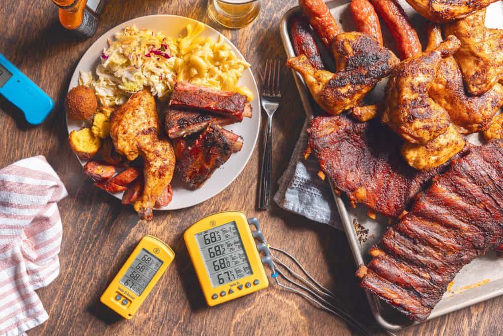 Platter of three meats, with a plate of meats and BBQ side dishes. Thermometer on the table. 