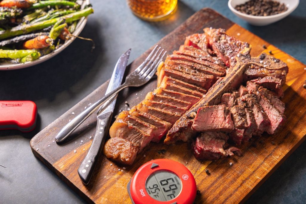 Porterhouse, sliced, on a cutting board with thermometers and charred vegetables