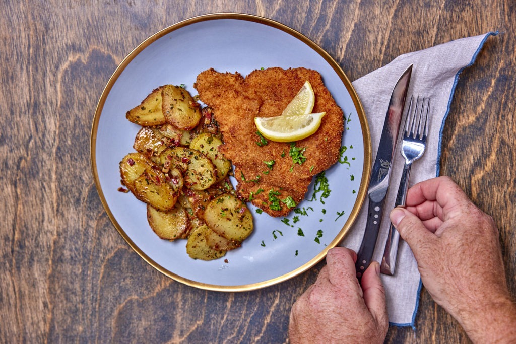 A plate of schnitzel and potatoes, hands setting a fork and knife next to it